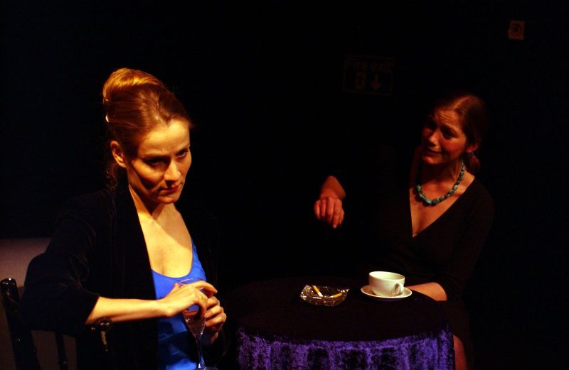 Two women (Amelia and Madam) are discussing across a round table with a dark blue velvet tablecloth, a single coffee cup and an ashtray.