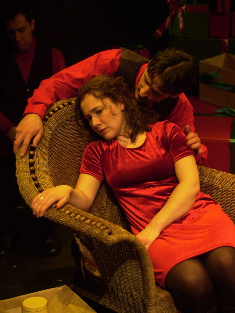 Nora is sitting in a wicker chair. Her facial expression and body language give the impression that she is sad and burdened by something. Helmer is standing behind the chair and is trying to make contact.