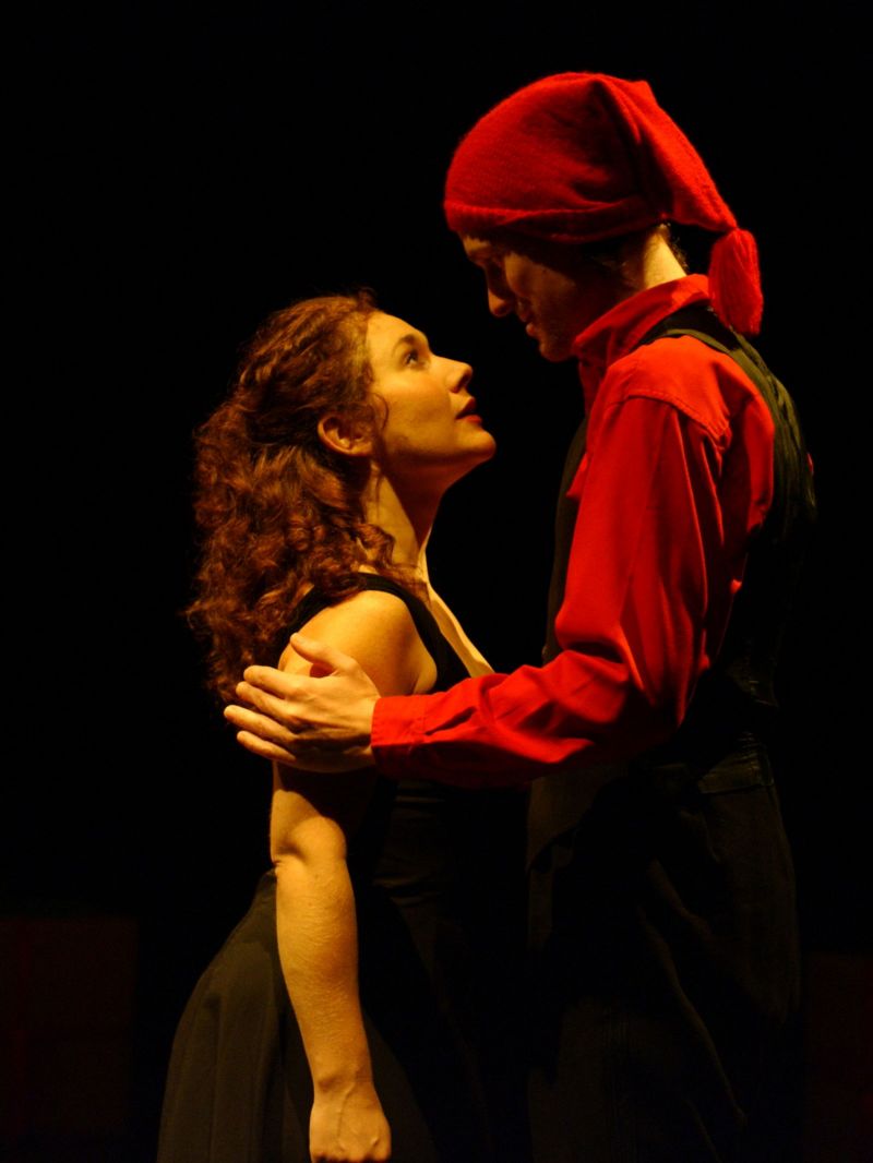 Nora and Helmer are standing upright looking at each other. Helmer is wearing a knitted woolly hat with a pom pom and is his hands on her upper arms looking her straight in the face suggesting he is reassuring or explaing her something.