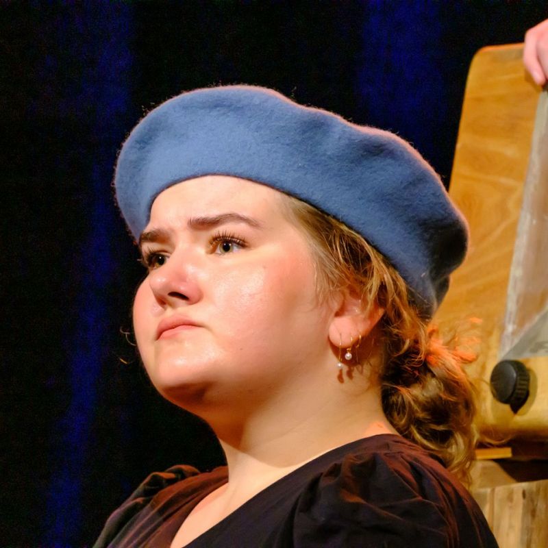 A head shot of a young woman with a blue beret looking ahead. her facial expression is seriuous and contemplative.
