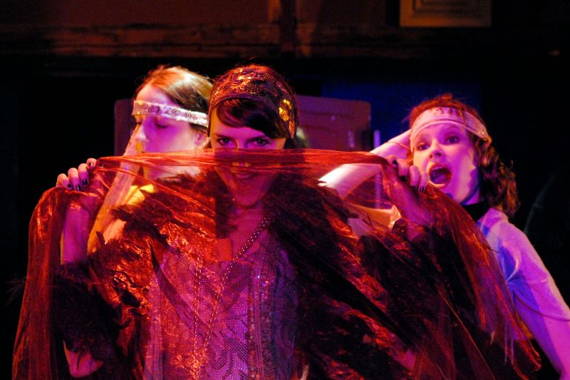 Three club girls in a speakeasy are performing. their act. The girl at the front hides half her face behind a red gauze.