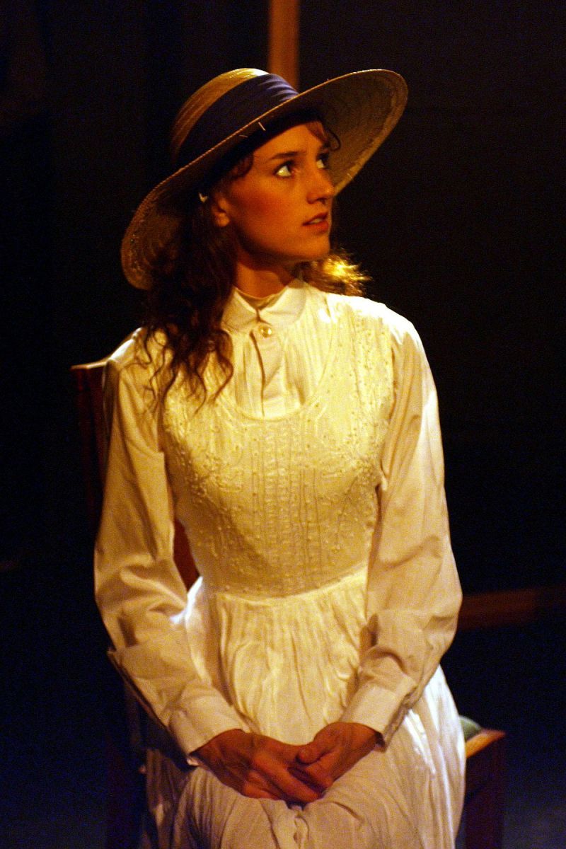 Dina Dorf is sitting on a bench. She is wearing a white summer dress and a straw hat with a blue ribbon. She is looking up at someone (whom one can&#039;t see).