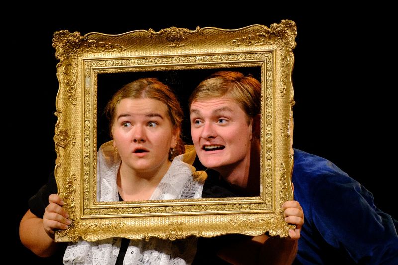 A young woman and a young man are looking through a small golden and elaborate period picture frame.