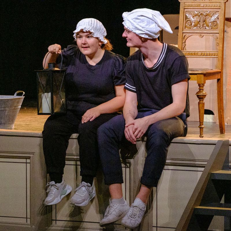 A young woman holding a lantern and a young man sitting on the edge of the stage. Both are wearing night caps.