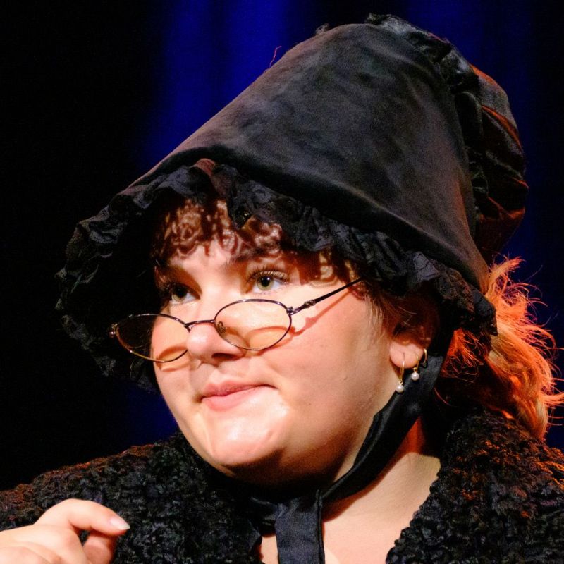 A head shot of a stern lady with glasses and a black bonnet. Her appearance and facial expression imply the suffragette movement