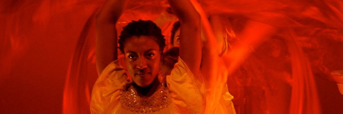 Anitra and a harem of girls are perfoming a dance with their veils barefoot. The lighting is a mix of amber and red.