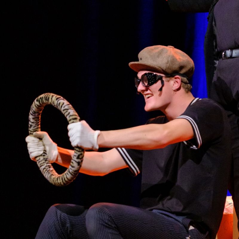 A young man wearing driving googles and a sixpence is holding onto a steering wheel pretending to go at fast speed.