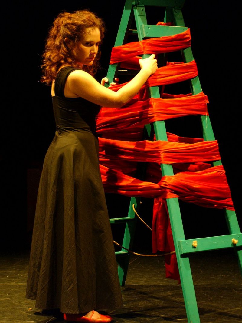 Nora is standing behind a step ladder wrapped in a long piece of red satin suggesting a Christmas tree. He facial expression is expressing contempation and a moment on her own in which she may be listening to a conversation in secret.