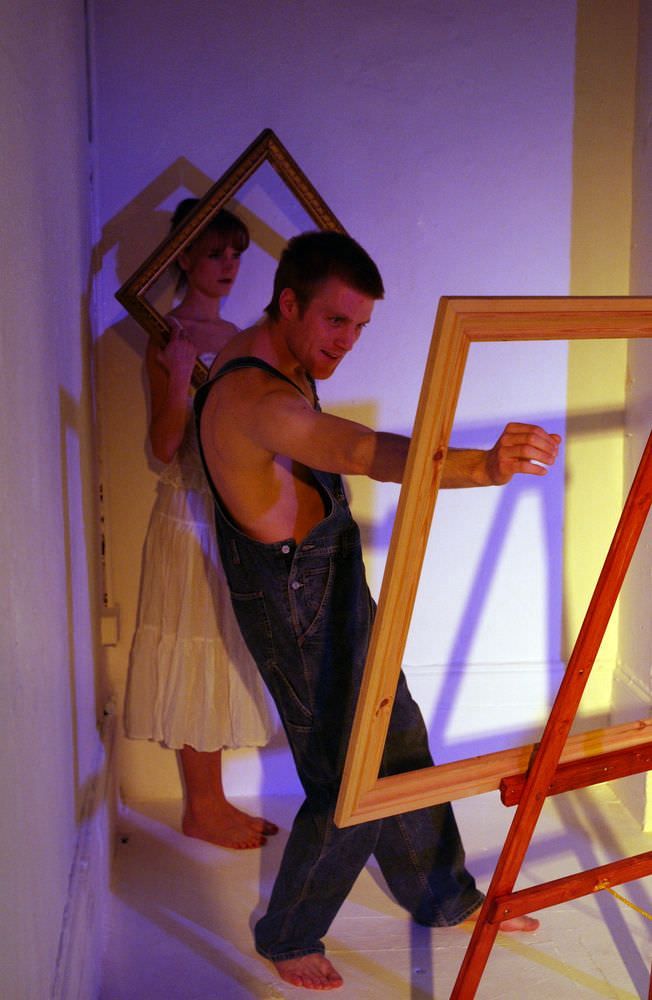 Axel is barefooted, barechested wearing an overall. He is working intensely on a painting in an empty picture frame.