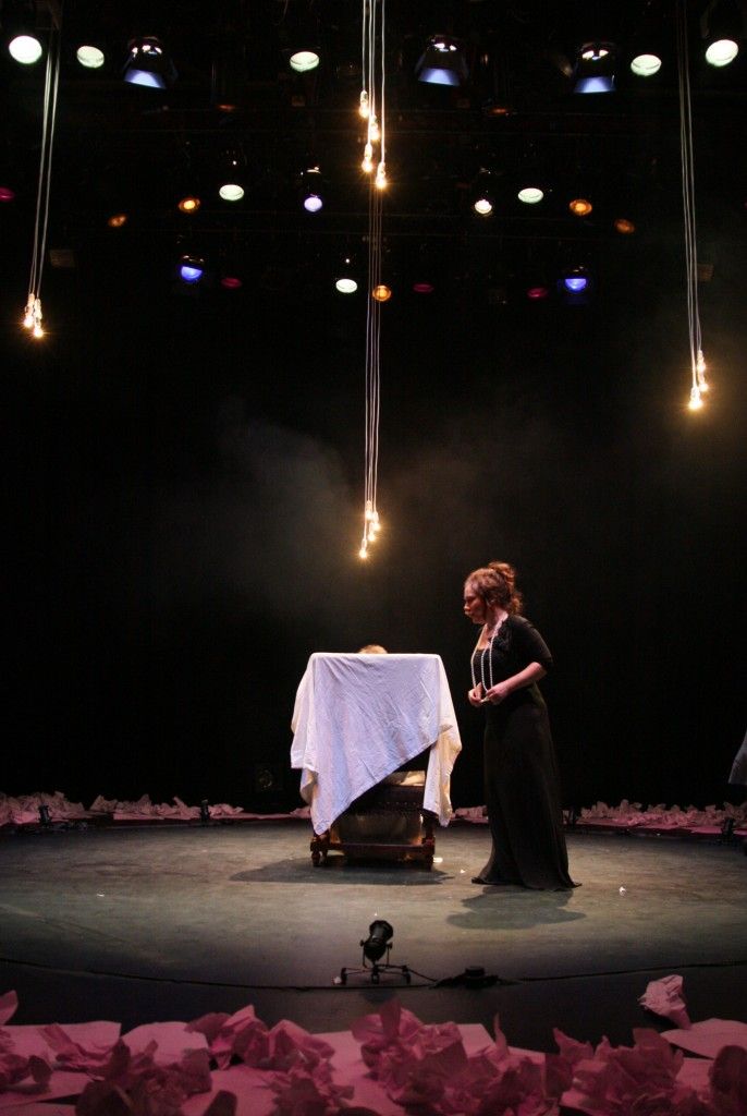 Hedda is walking around in an empty circle surrounded by crushed sheets of paper. lit in purple. Her body language gives the impression of a caged animal.